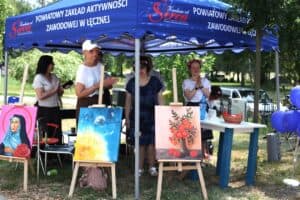 Niebieski namiot Powiatowego Zakłądu Aktywności Zawodowej w Łęcznej „Kuchnia od Serca” z osobami w środku. Na zewnątrz, na sztalugach ustawiono obrazy przedstawiające m.in. postać w błękitnym szalu oraz bukiet kwiatów.
