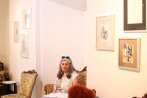 Sonia Wilk siedzi w zabytkowym fotelu i patrzy w bok. Na ścianie za nią wiszą oprawione rysunki postaci.
