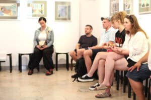 Grupa osób siedzi rzędem na taboretach wzdłuż ściany galerii, na której wiszą obrazy. Uczestnicy są zwróceni w jedną stronę.