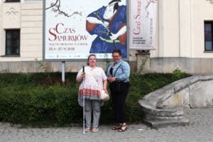 Dwie kobiety, w tym jedna z białą laską, stoją przed budynkiem Muzeum Narodowego w Lublinie. Obok znajduje się billboard reklamujący wystawę „Czas Samurajów” z ilustracją japońskiego wojownika.