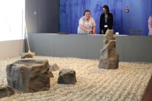 Kobieta z białą laską i pracowniczka muzeum stoją przy pulpicie interaktywnym. Przed nimi znajduje się ogród kamienny.