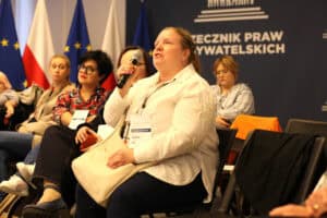 Ewelina Orzechowska mówi do mikrofonu podczas spotkania. W tle widać pozostałych uczestników spotkania oraz flagi Polski i Unii Europejskiej.