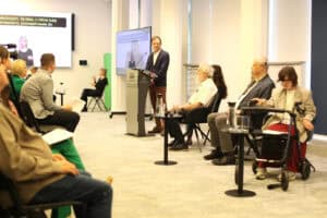 Adam Krzywoń stoi przy mównicy i mówi do mikrofonu. Obok siedzą pozostali paneliści, w tym kobieta z balkonikiem rehabilitacyjnym. Na ekranie wyświetlany jest tłumacz języka migowego.