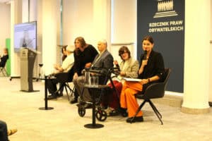Panel dyskusyjny w Biurze Rzecznika Praw Obywatelskich. Kobieta w czarnej marynarce i pomarańczowych spodniach mówi do mikrofonu. Obok siedzą pozostali paneliści, w tym osoba z balkonikiem rehabilitacyjnym.