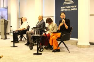 Panel dyskusyjny w Biurze Rzecznika Praw Obywatelskich. Kobieta w czarnej marynarce i pomarańczowych spodniach mówi do mikrofonu i gestykuluje. Obok siedzą pozostali paneliści.