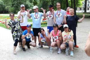 Grupa uczestników z medalami pozuje do wspólnego zdjęcia w parku i pokazuje gesty radości.