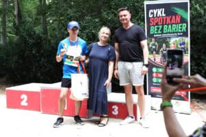 Uczestnik biegu z medalem i dyplomem stoi przy podium z dwiema osobami.