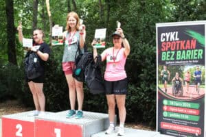 Trzy uczestniczki stoją na podium z medalami, pucharami i dyplomami. Obok stoi baner wydarzenia „Cykl Spotkań Bez Barier”.