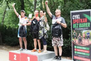 Uczestnicy biegu stoją na podium z medalami, pucharami i dyplomami. Wszyscy unoszą ręce.