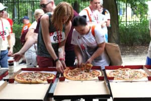 Dwie kobiety w strojach sportowych z medalami sięgają po kawałki pizzy z pudełek na stole.