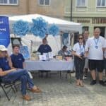 Zadaszone białe stoisko Warsztatu Terapii Zajęciowej (WTZ) przy Fundacji Centrum Niepełnosprawnych Przedsiębiorców (FCNP), ozdobione błękitnymi kwiatami, stoi na placu miejskim w Chełmie. Pod zadaszeniem oraz obok stoiska uczestnicy i instruktorzy WTZ pozują do wspólnego zdjęcia.