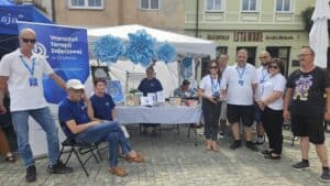 Zadaszone białe stoisko Warsztatu Terapii Zajęciowej (WTZ) przy Fundacji Centrum Niepełnosprawnych Przedsiębiorców (FCNP), ozdobione błękitnymi kwiatami, stoi na placu miejskim w Chełmie. Pod zadaszeniem oraz obok stoiska uczestnicy i instruktorzy WTZ pozują do wspólnego zdjęcia.