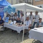 Uczestnicy i instruktorzy Warsztatu Terapii Zajęciowej przy Fundacji Centrum Niepełnosprawnych Przedsiębiorców pozują do zdjęcia podczas wydarzenia na placu miejskim w Chełmie. Obok znajduje się stoisko fundacji z rollupem i materiałami dla uczestników.