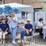 Od lewej stoją instruktor Tomasz Karenko i kierownik Warsztatu Terapii Zajęciowej Agnieszka Gruszecka-Sędłak, a siedzą uczestnicy WTZ Anna Karaś oraz uczestnik Warsztatu. Tomasz Karenko i Agnieszka Gruszecka-Sędłak układają dłonie w kształt serca. Przy sąsiednim stole Fundacji Centrum Niepełnosprawnych Przedsiębiorców siedzą instruktorzy i dzieci podczas wydarzenia na placu miejskim w Chełmie.