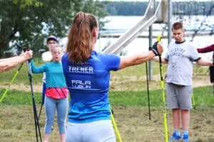 Anna Majerska w niebieskiej koszulce z napisem „Trener” na plecach, prowadzi rozgrzewkę z kijkami do Nordic Walking dla grupy młodzieży nad jeziorem.