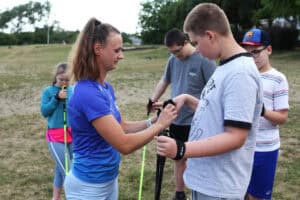 Anna Majerska pomaga chłopcu dopasować rękawiczki i paski do kijków Nordic Walking. W tle grupka młodzieży.