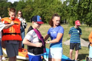 Instruktorka zapina kamizelkę asekuracyjną jednemu z uczestników. W tle grupa osób i kolorowe kajaki.
