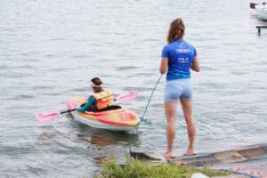 Trenerka stoi na brzegu jeziora i trzyma linę asekuracyjną, doczepioną do małego, kolorowego kajaka, w którym siedzi dziewczynka.