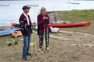 Kobieta i mężczyzna stoją nad brzegiem zalewu z kijkami do Nordic Walking. Za nimi leżą kajaki.