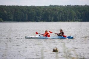 Dwie osoby płyną kajakiem po jeziorze. Za nimi rozciąga się zalesiony brzeg.