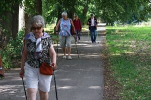 Grupa seniorów spaceruje z kijami do Nordic Walking po asfaltowej ścieżce między drzewami.