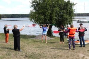 Grupa osób z instruktorką wykonuje rozgrzewkę nad brzegiem zalewu.