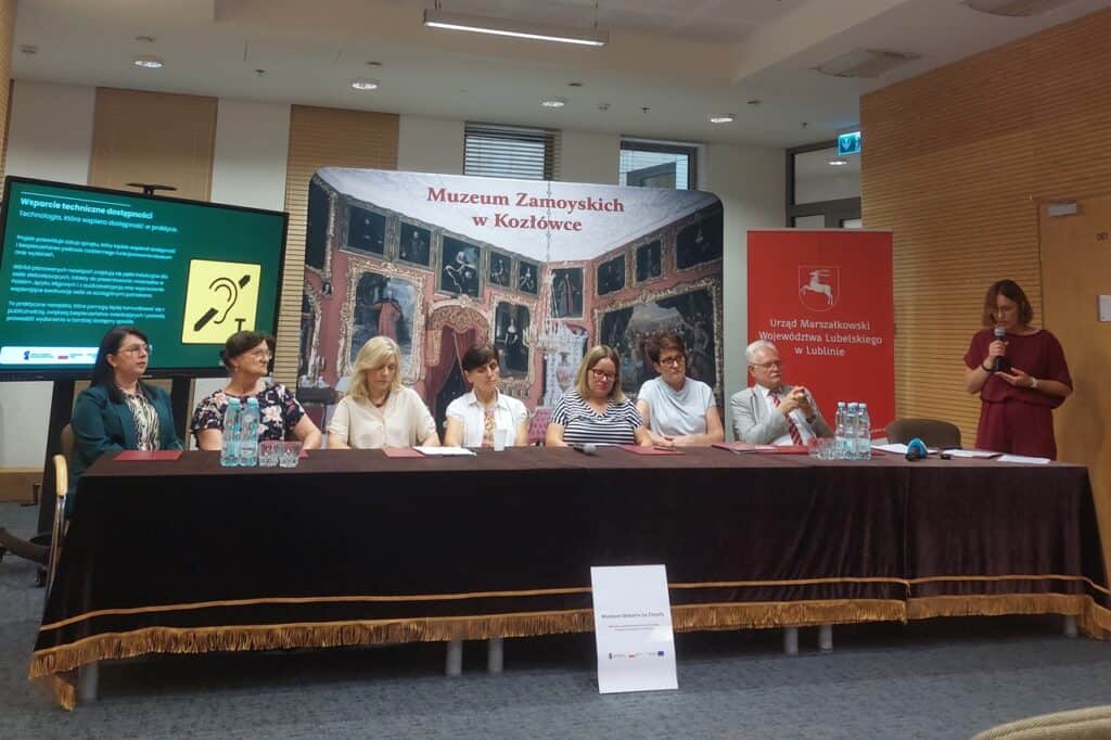 Przy stole konferencyjnym siedzi siedmioro reprezentantów instytucji i organizacji współpracujących przy realizacji projektu, po prawej stoi prowadząca z mikrofonem; w tle banery Muzeum Zamoyskich w Kozłówce oraz Urzędu Marszałkowskiego Województwa Lubelskiego i ekran z prezentacją.
