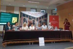 Przy stole konferencyjnym siedzi siedmioro reprezentantów instytucji i organizacji współpracujących przy realizacji projektu, po prawej stoi prowadząca z mikrofonem; w tle banery Muzeum Zamoyskich w Kozłówce oraz Urzędu Marszałkowskiego Województwa Lubelskiego i ekran z prezentacją.