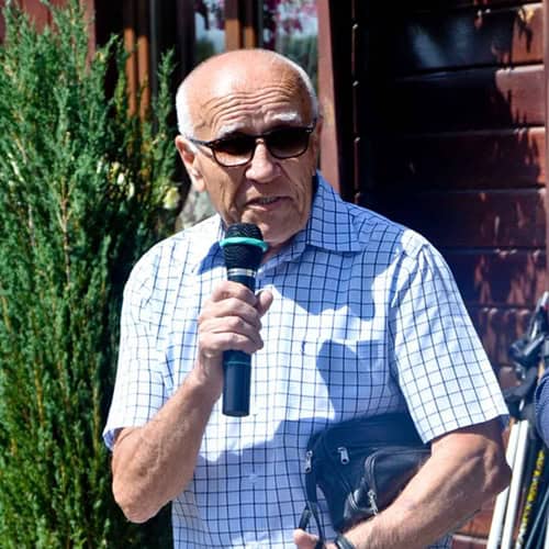 Kazimierz Telega, siwy mężczyzna w okularach, stoi w słoneczny dzień przed budynkiem restauracji Zacisze w Międzyrzecu Podlaskim. Trzyma w dłoni mikrofon, a w tle widoczna jest elewacja budynku; fot. Hubert Juszczuk