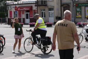 Kobieta w zielonej koszulce macha ręką do uczestników. Przed nią siedzi mężczyzna na rowerze. Po lewej stoi odwrócony tyłem strażak.