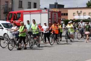 Grupa rowerzystów w kamizelkach odblaskowych przejeżdża przez plac miejski. W tle czerwony wóz strażacki i zabudowania.