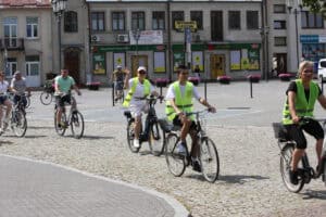 Uczestnicy rajdu rowerowego jadą przez brukowany plac miejski na tle kamienic i sklepów.