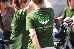 Plecy kobiety w zielonej koszulce z widocznym logo fundacji „PoMOC Daje MOC”.