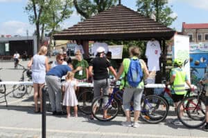 Grupa osób z rowerami stoi przy stoliku pod drewnianą altaną na placu miejskim. W tle zabudowania.
