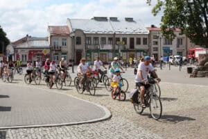 Kolumna uczestników rajdu w różnym wieku porusza się brukowaną ulicą wokół miejskiego placu.