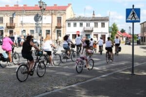 Grupa rowerzystów jedzie ulicą w kierunku ulicy zabudowanej kamienicami. Po prawej stronie znak przejścia dla pieszych.