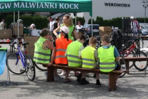 Dzieci i dorośli w kamizelkach odblaskowych siedzą na drewnianej ławce. W tle stoisko KGW Malwianki Kock.