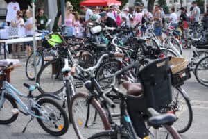 Rzędy rowerów zaparkowanych na placu. W tle grupa osób w różnym wieku.