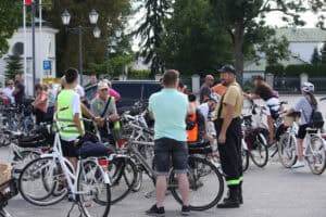 Grupa mężczyzn rozmawia na placu wśród zaparkowanych rowerów. W tle inne osoby i zieleń drzew.