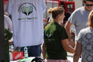 Na stoisku wyeksponowano okolicznościową koszulkę z logo Fundacji „PoMOC Daje MOC”. Obok stoi odwrócona tyłem kobieta w zielonej koszulce z logo fundacji.