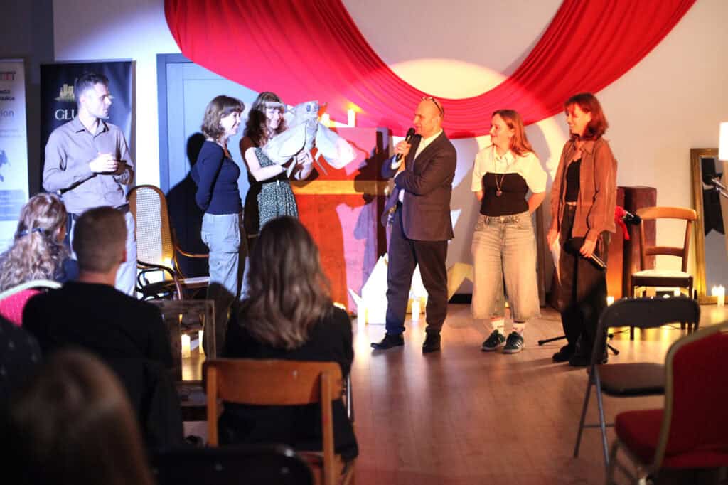 Sześć osób stoi na scenie przed publicznością. Mężczyzna w marynarce mówi do mikrofonu, kobieta trzyma rekwizyt mola, a tłumacz języka migowego tłumaczy wystąpienie.