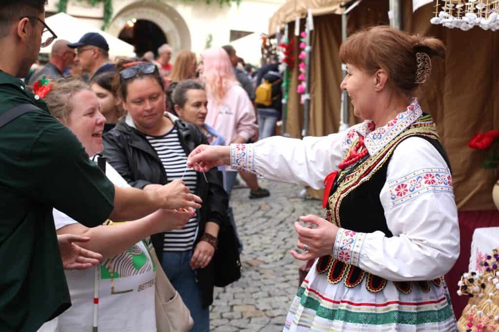 Kobieta w bogato zdobionym stroju ludowym wręcza drobny przedmiot kobiecie przy stoisku jarmarcznym. Obok stoją inne osoby, w tym kobieta w bluzce w paski.