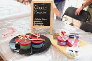 Na stole stoją gotowe świece w różnych kolorach i formach. Obok znajduje się tabliczka z napisem „Czucie Poczucia”.