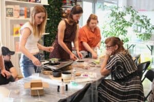 Grupa kobiet przy wspólnym stole. Dwie obsługują przenośną kuchenkę elektryczną z garnkiem do topienia wosku. Inne kobiety obserwują proces. Na stole widać pojemniki na wosk, olejki eteryczne i formy.