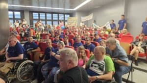 Uczestnicy II Festiwalu „Chełm śpiewa bez barier” siedzą na widowni. Wśród nich są osoby w niebieskich koszulkach, osoby na wózkach oraz opiekunowie. W tle uczestnicy trzymają transparenty.