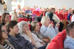 Na widowni siedzi publiczność, w tym organizatorzy i zaproszeni goście. Uśmiechają się, śmieją i wskazują w stronę sceny.