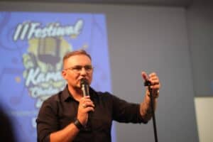 Wokalista zespołu „Nokaut” występuje na scenie podczas II Festiwalu „Chełm śpiewa bez barier”. Trzyma mikrofon i stojak na mikrofon. W tle logo wydarzenia.