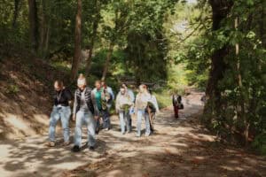 Grupa kobiet spaceruje piaszczystą ścieżką wśród drzew. Przez korony drzew prześwieca słońce.