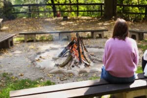 Kobieta w różowym swetrze siedzi tyłem na drewnianej ławce i patrzy na ognisko w kamiennym kręgu. Wokół znajduje się jesienny las i puste ławki.