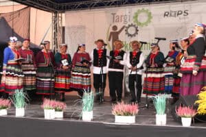 Kobiety i mężczyźni w tradycyjnych, kolorowych strojach ludowych stoją na scenie i śpiewają do mikrofonów. Za nimi znajduje się baner „Targi Ekonomii Społecznej ROPS Lublin”.